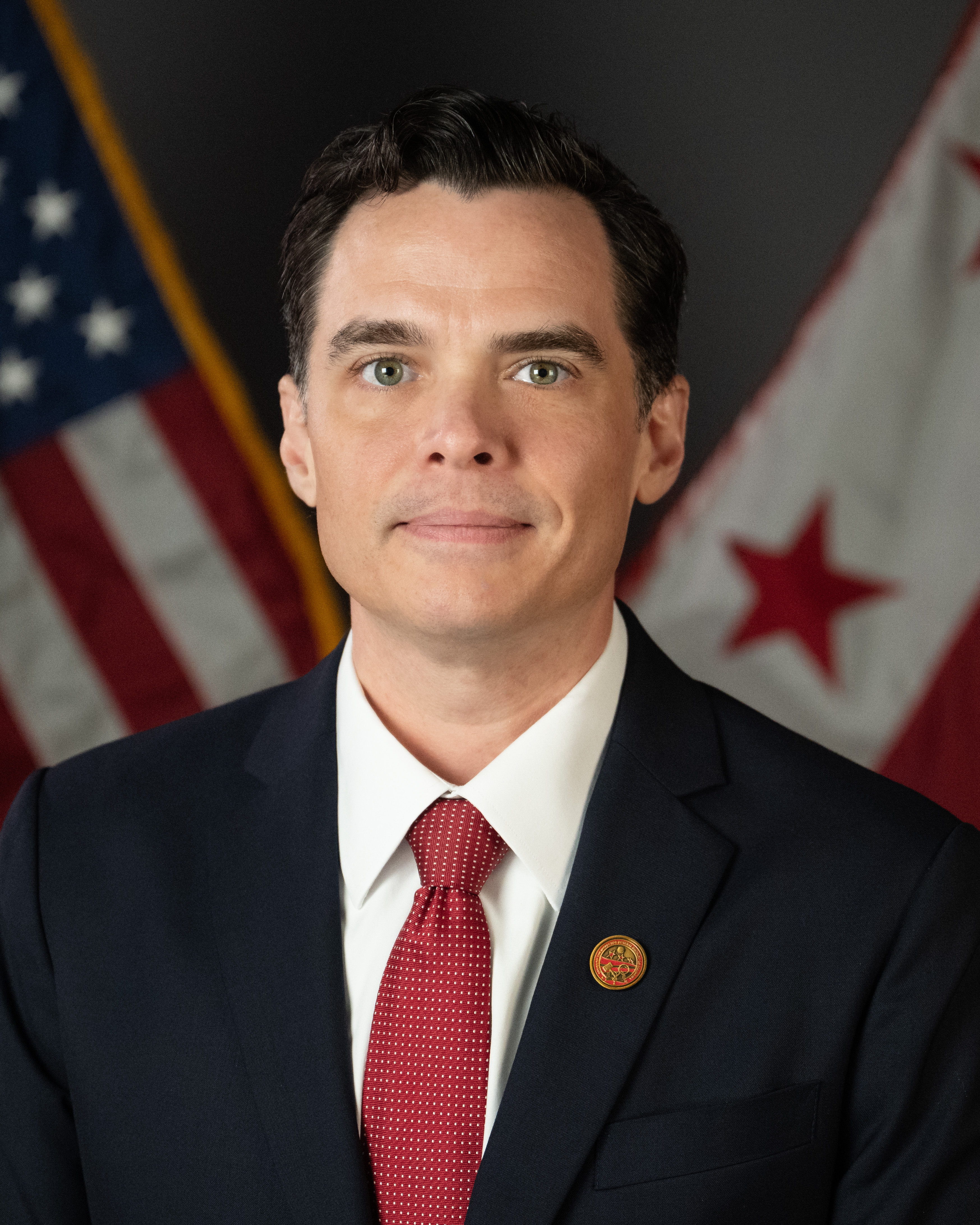 Upper‑torso portrait of an individual wearing a navy suit jacket, white dress shirt, and red textured tie. A circular red‑and‑gold lapel pin appears on the left lapel. The US flag (stars and stripes) is positioned to the left; the District of Columbia flag (three red stars above two red bars) is positioned to the right. Neutral dark studio backdrop with even lighting; subject centered with shoulders slightly angled.